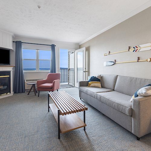 A modern living room features a gray sofa, a pink chair, a fireplace, a TV, and a coffee table, with balcony access and ocean views ending.