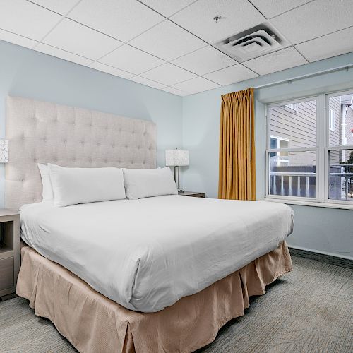 A neatly arranged bedroom with a large bed, nightstands, lamps, and a window with yellow curtains. The space is modern with soft, neutral colors.