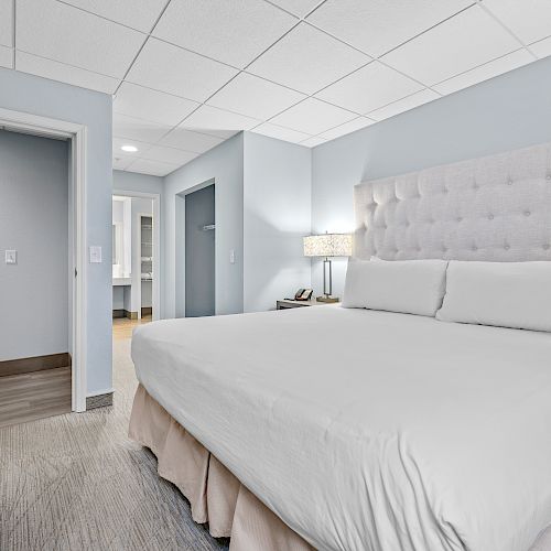 A modern hotel room with a large bed, white bedding, nightstand with a lamp, dresser, and open doorways leading to the bathroom and hallway.