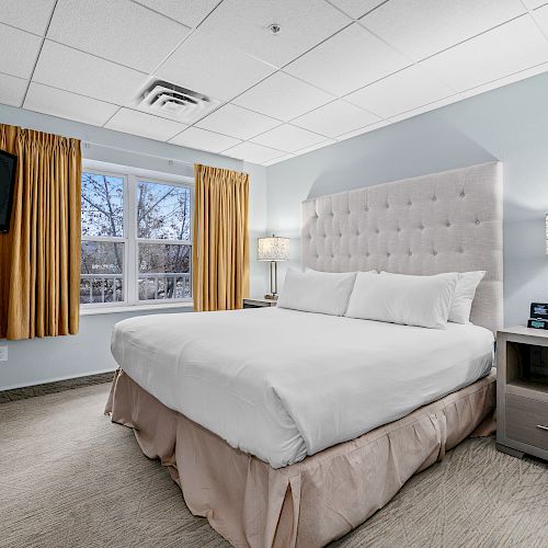 The image shows a neatly made bed in a hotel room with a padded headboard, two bedside tables, a wall-mounted TV, and a window with yellow curtains.