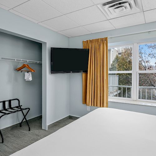 A room with a TV, a closet with a hanging rod and suitcase rack, and a window with yellow curtains, providing a view of trees outside.