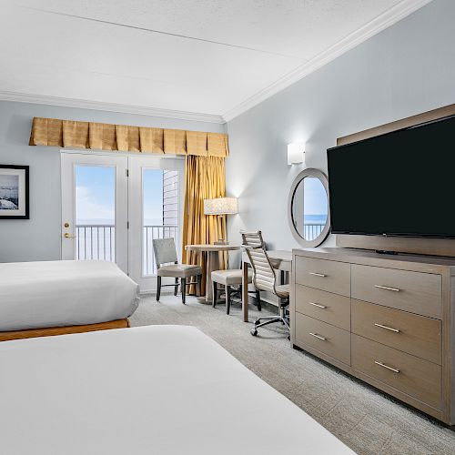 A hotel room with two beds, a TV on a dresser, a desk and chair, a balcony with ocean view, and a framed picture on the wall.