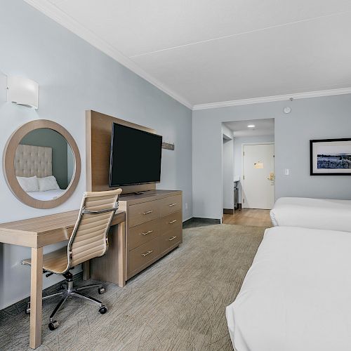 The image shows a hotel room with two beds, a TV on a dresser, a desk with a chair, a wall mirror, and an exit door.