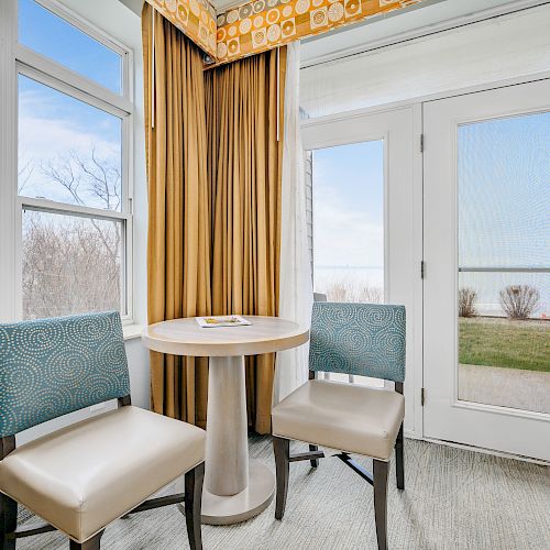 The image shows a small seating area with two chairs and a round table by windows and a glass door with a view of trees and water.