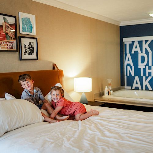 Two children on a bed in a cozy room with framed art on the wall and a jacuzzi tub in the background that has a sign saying 