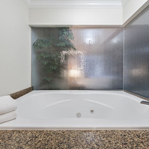 This image shows a bathroom with a white bathtub, folded towels on the edge, and a textured glass window with a plant visible through it.