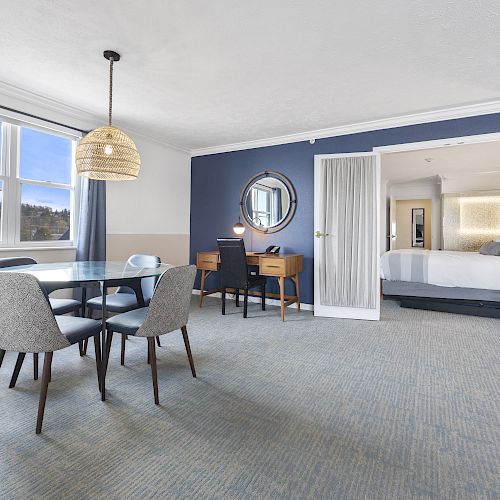 A spacious hotel room with a seating area, desk, round mirror, and a bedroom with a large bed. The room has blue and white decor.