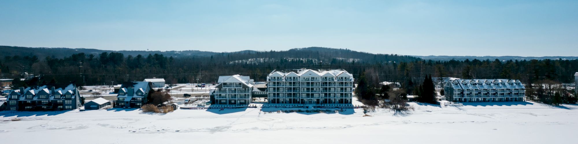 A row of buildings stands along a snowy landscape next to a partially frozen body of water, under a clear blue sky.