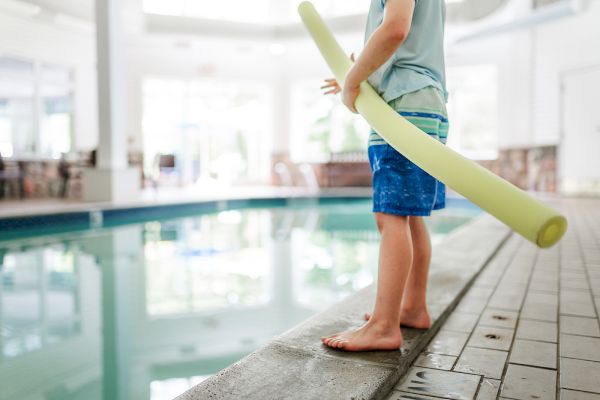 A child in swim shorts stands at the edge of an indoor pool, holding a yellow pool noodle, preparing to enter the water.