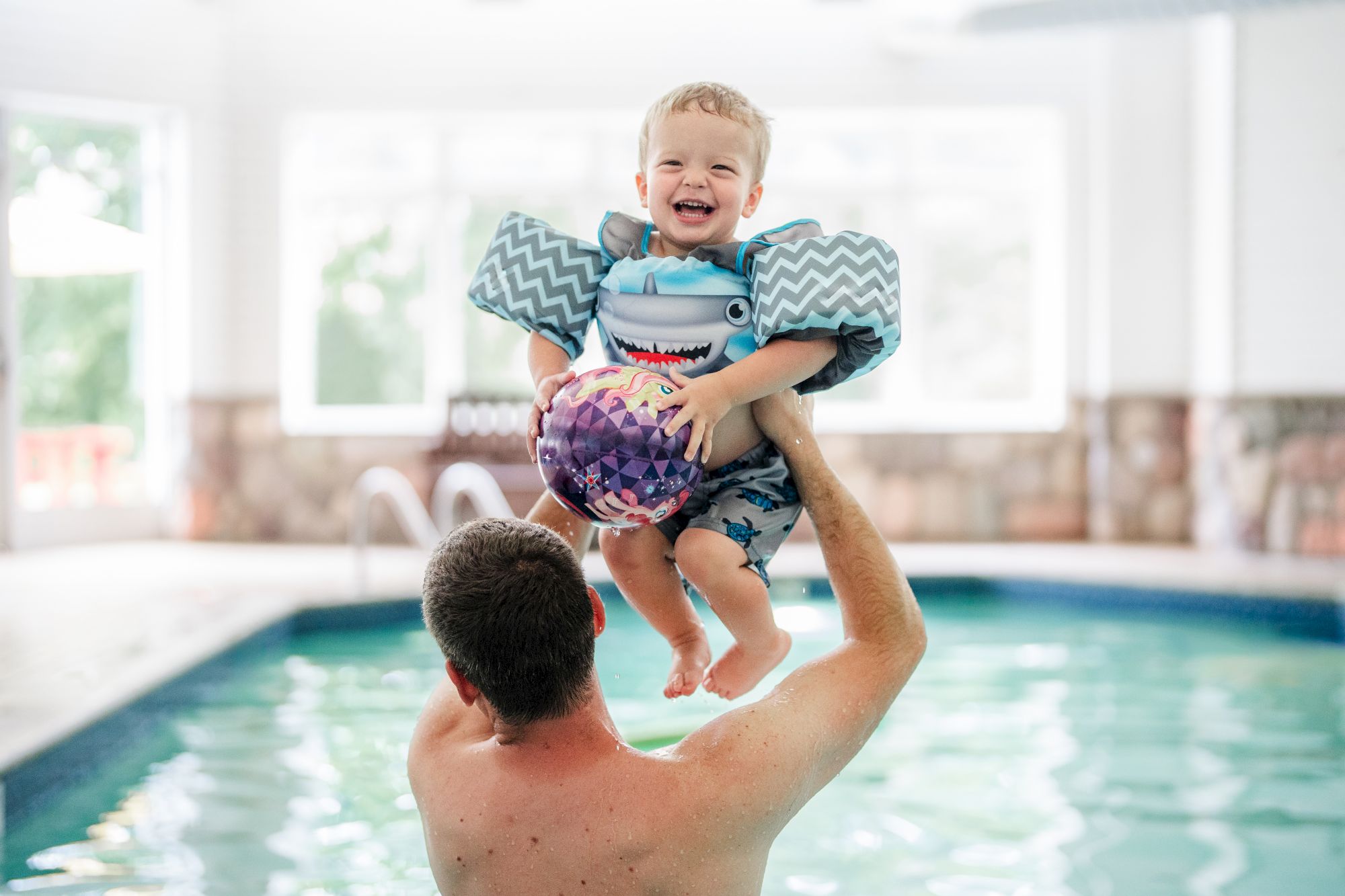 An adult lifts a laughing child wearing arm floaties and holding a ball in an indoor pool.