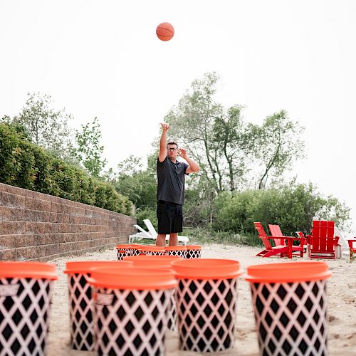 A person is playing a beach basketball game, throwing a ball towards large buckets. Red chairs and greenery are in the background.