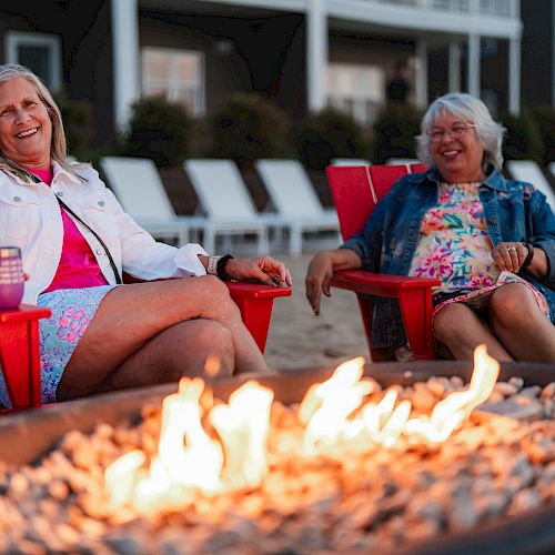 Two women in colorful outfits sit in red chairs by a fire pit, smiling and enjoying their time, with a backdrop of lounge chairs and a building.