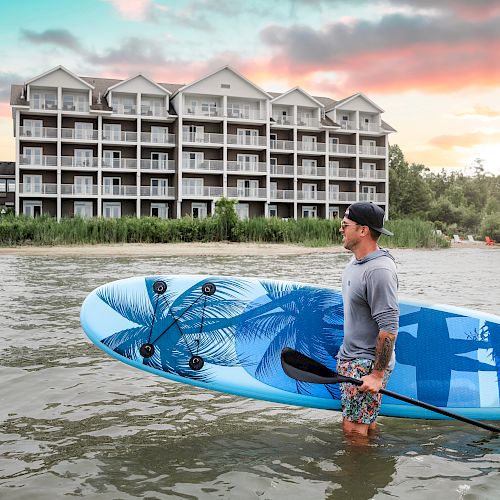 A man stands in the water holding a paddleboard in front of a large building with multiple balconies, against a sunset sky with clouds.