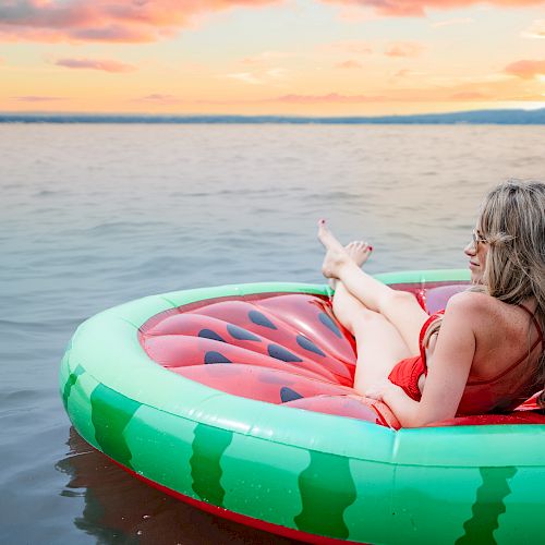 A person relaxes on a watermelon-themed float in a body of water during sunset, enjoying a calm and serene moment.