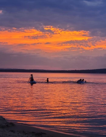 People are enjoying water activities on a calm lake during a colorful sunset, with a beautiful orange and purple sky reflecting in the water.