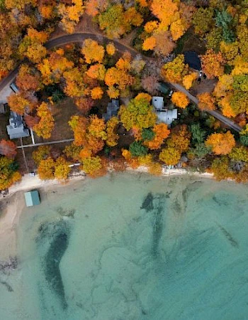 Aerial view of a lakeside area with autumn trees, a beach, tennis courts, and nearby buildings surrounded by vibrant fall foliage.