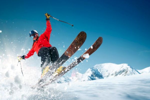 A skier in a red jacket is performing a jump on snowy slopes with mountains in the background under a clear blue sky.