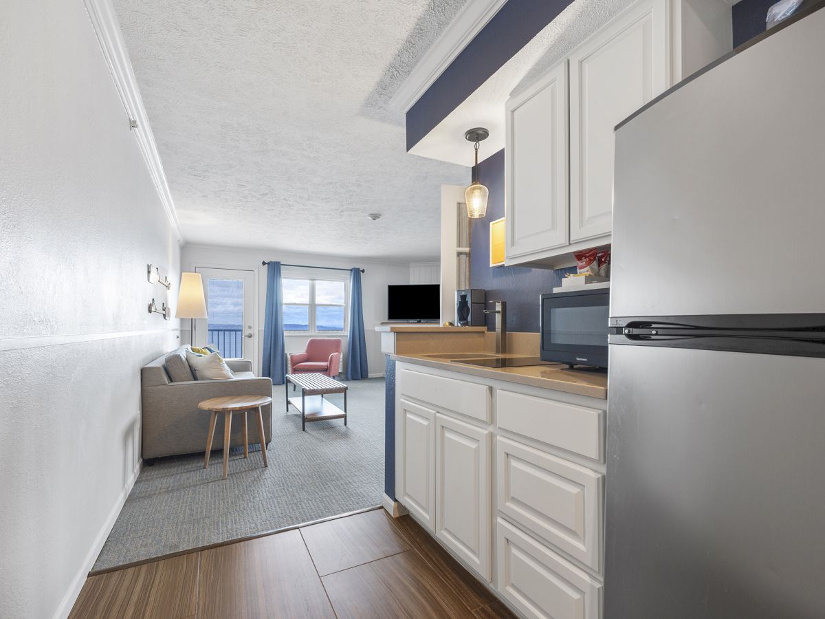 The image shows a modern apartment interior with a small kitchen, living area, couch, TV, and natural light from large windows.