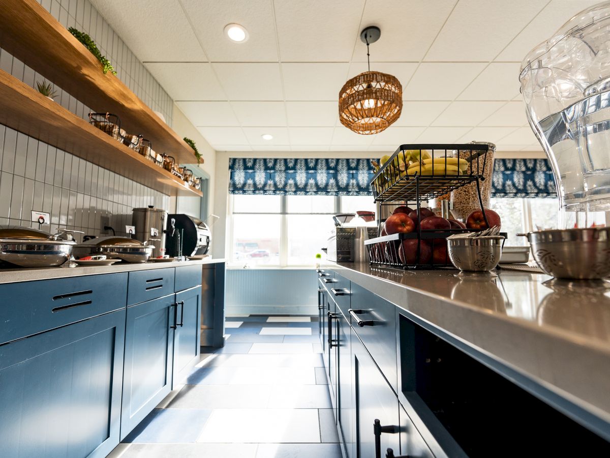 A modern kitchen with blue cabinets, open shelves, fruit baskets, and a ceiling lamp, viewed from a low angle facing the window.