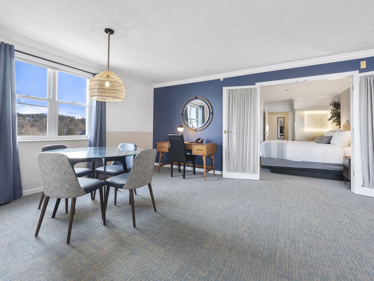 The image shows a modern hotel room with a dining area, desk, and bed. Curtains, a pendant light, and a wall mirror add to the decor.