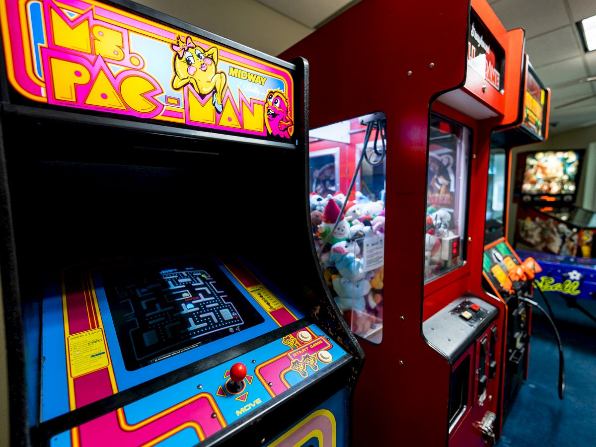 The image shows an arcade with a Ms. Pac-Man machine, a claw machine with plush toys, and other arcade games in the background.