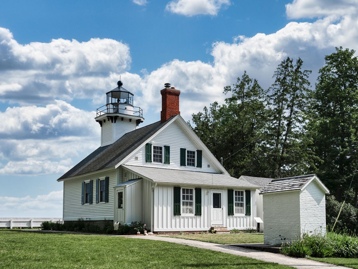 A quaint white lighthouse with a red chimney is framed by green trees and a cloudy blue sky, sitting on a grassy landscape.