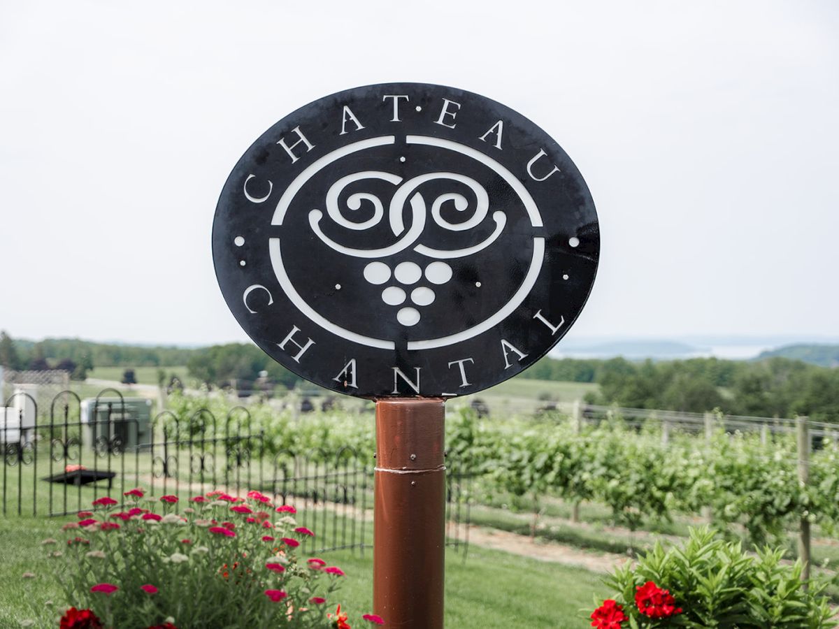 A "Chateau Chantal" sign stands in front of vineyards with some red flowers in the foreground and a scenic view in the background.