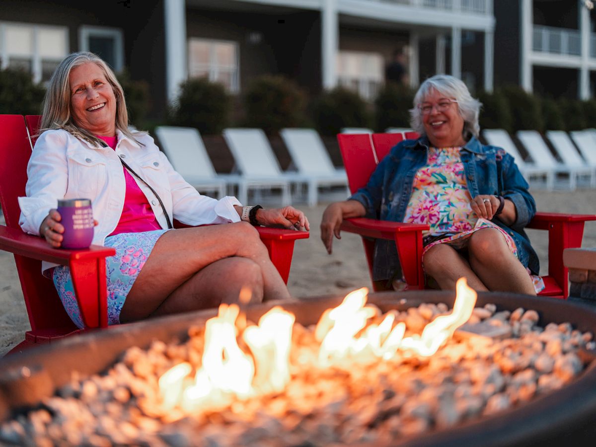 Two people are smiling and relaxing in red chairs by a fire pit, with lounge chairs and a building in the background.