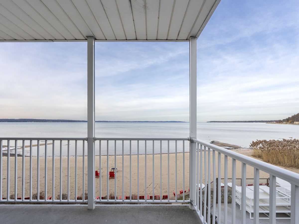 A view from a covered balcony overlooking a sandy beach and calm sea, with clear skies and distant shoreline.