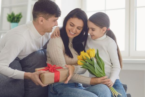 A family is sitting on a couch. The man hands a gift to the woman, while the girl offers her yellow tulips. They're joyful together.