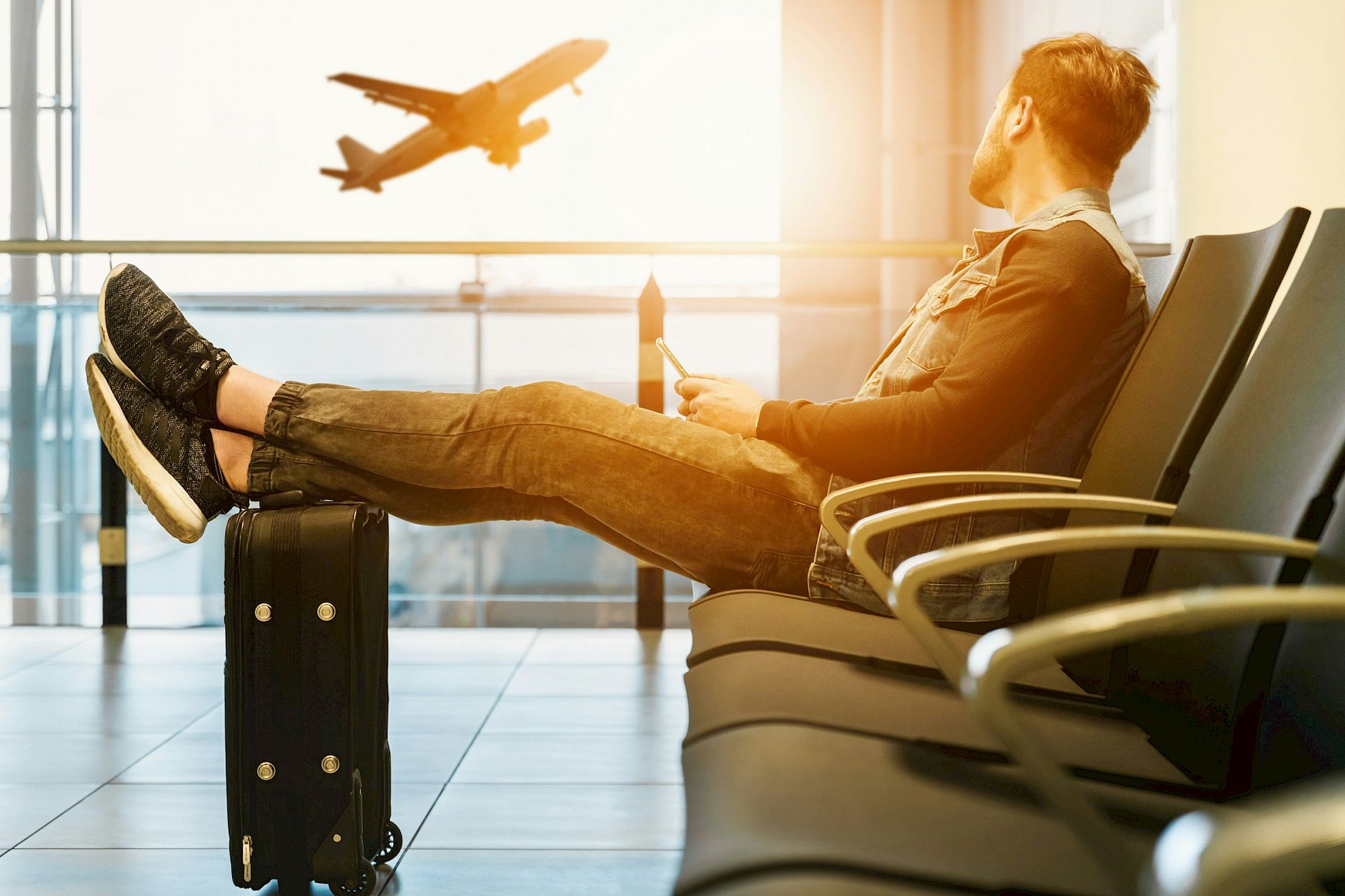A person sits in an airport lounge with feet on a suitcase, watching a plane take off through the window.