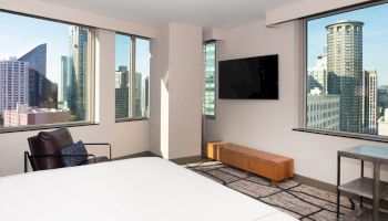 A modern hotel room features floor-to-ceiling windows with a cityscape view, a wall-mounted TV, a bed, a chair, and minimalist furnishings.