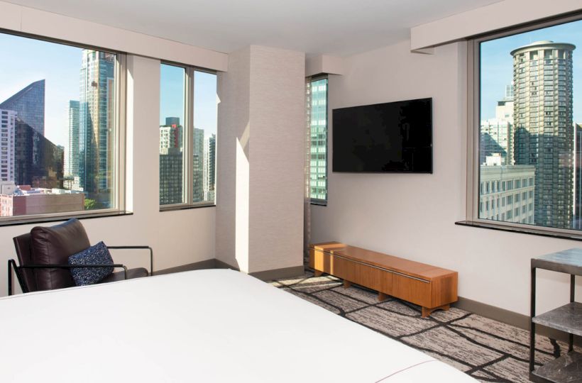 A modern hotel room features floor-to-ceiling windows with a cityscape view, a wall-mounted TV, a bed, a chair, and minimalist furnishings.