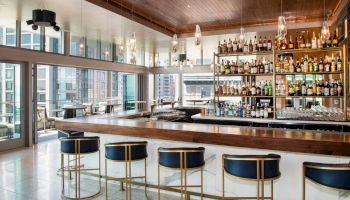 A modern bar with a wooden countertop, stylish bar stools, and shelves filled with bottles. Large windows provide views of a cityscape.