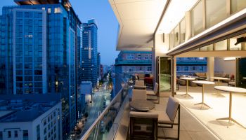 This image shows a modern rooftop terrace with tables and chairs, overlooking a cityscape with tall buildings at dusk or dawn.