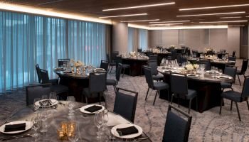 This image shows an elegant dining area set up for an event, with round tables, dark chairs, and place settings, including glasses and napkins.