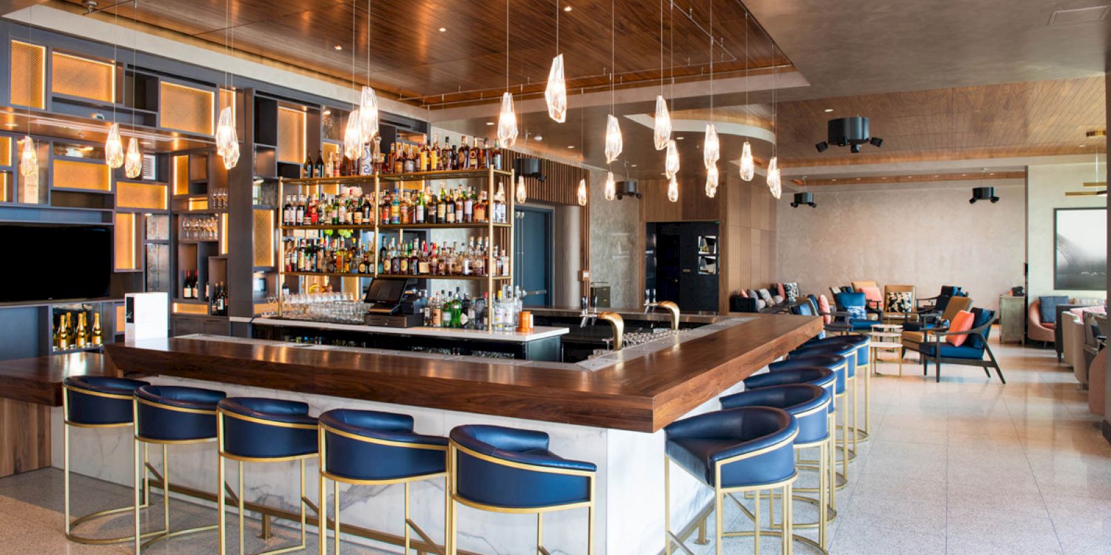 This is an image of a modern bar with a wooden counter, blue and gold bar stools, hanging lights, and a well-stocked liquor display behind the counter.