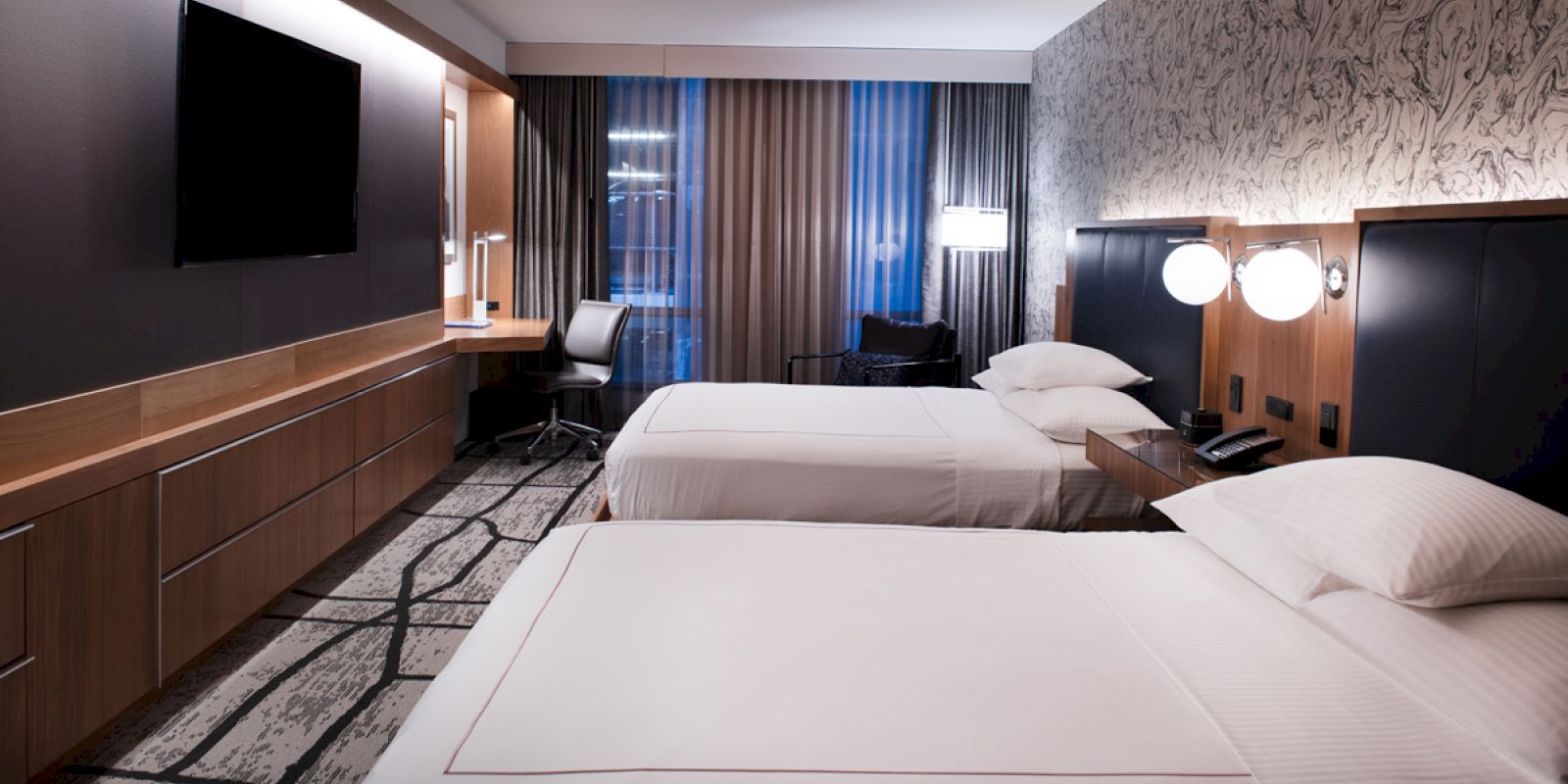 A modern hotel room with two beds, a TV, a desk, a chair, and contemporary decor.