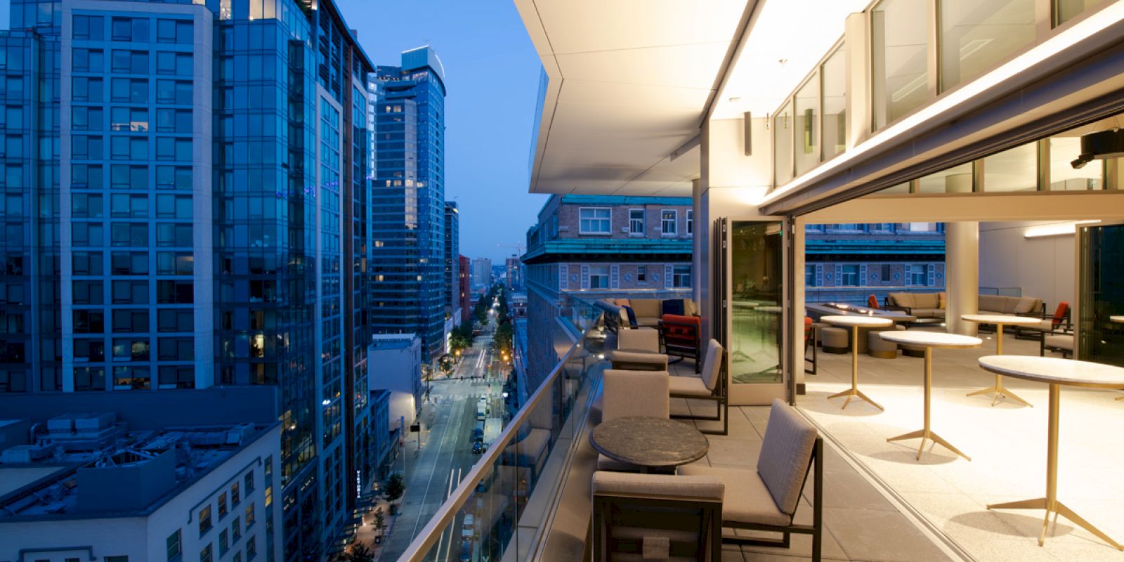 An image of a modern cityscape with high-rise buildings at dusk, viewed from a balcony with outdoor seating and tables.