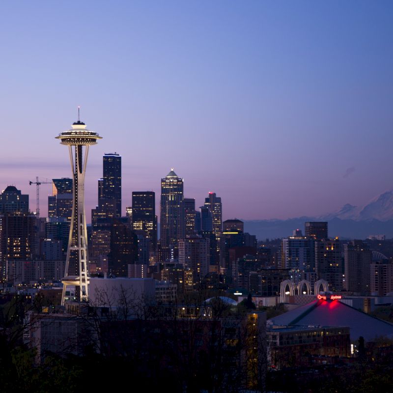 This image shows the Seattle skyline at dusk, featuring the Space Needle with Mount Rainier visible in the background, set under a twilight sky.
