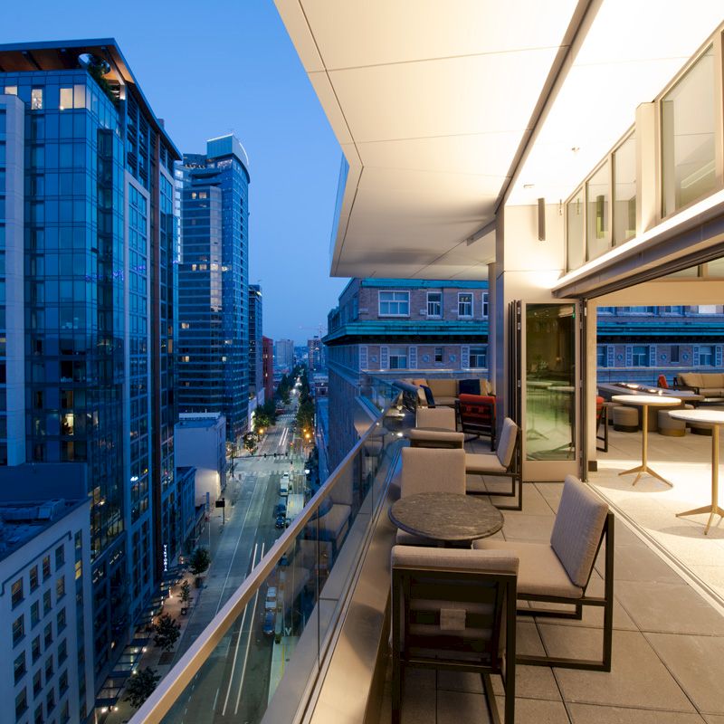 A modern balcony view overlooking a city street at dusk, with comfortable seating and tables, adjacent to large buildings and illuminated interiors.