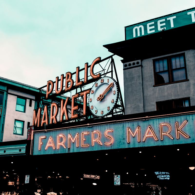 The image shows the entrance to a public market with the signs "PUBLIC MARKET" and "FARMERS MARKET" illuminated by neon lights, alongside other buildings.