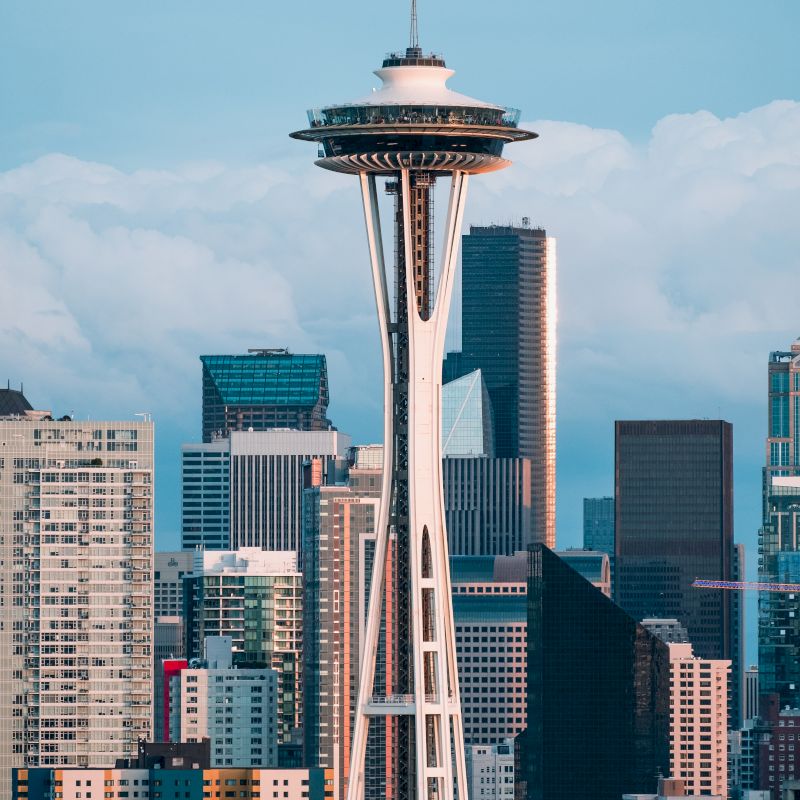The image shows the Seattle Space Needle with a backdrop of several modern skyscrapers and buildings under a partly cloudy sky.