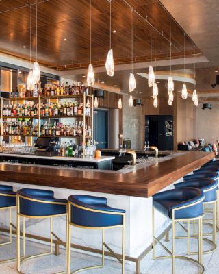 A modern bar with a wooden counter, high blue chairs, pendant lights, a fully stocked back shelf, and a lounge area with sofas and chairs.