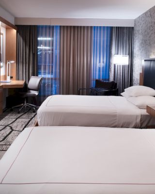 A modern hotel room with two neatly made beds, a wall-mounted TV, a desk with a chair, and large windows with curtains is shown.