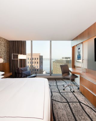 A modern hotel room features a large bed, a wall-mounted TV, a desk with a chair, a stylish chair near the window, and a view of buildings outside.