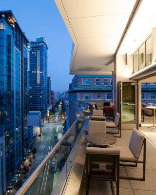 A modern city evening view from a high-rise balcony with lounge chairs and tables, looking out over the street and adjacent buildings.
