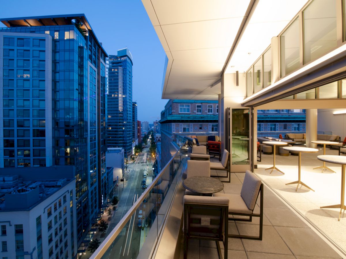 The image shows a modern cityscape during twilight with tall buildings and a sleek, open balcony featuring tables and chairs.