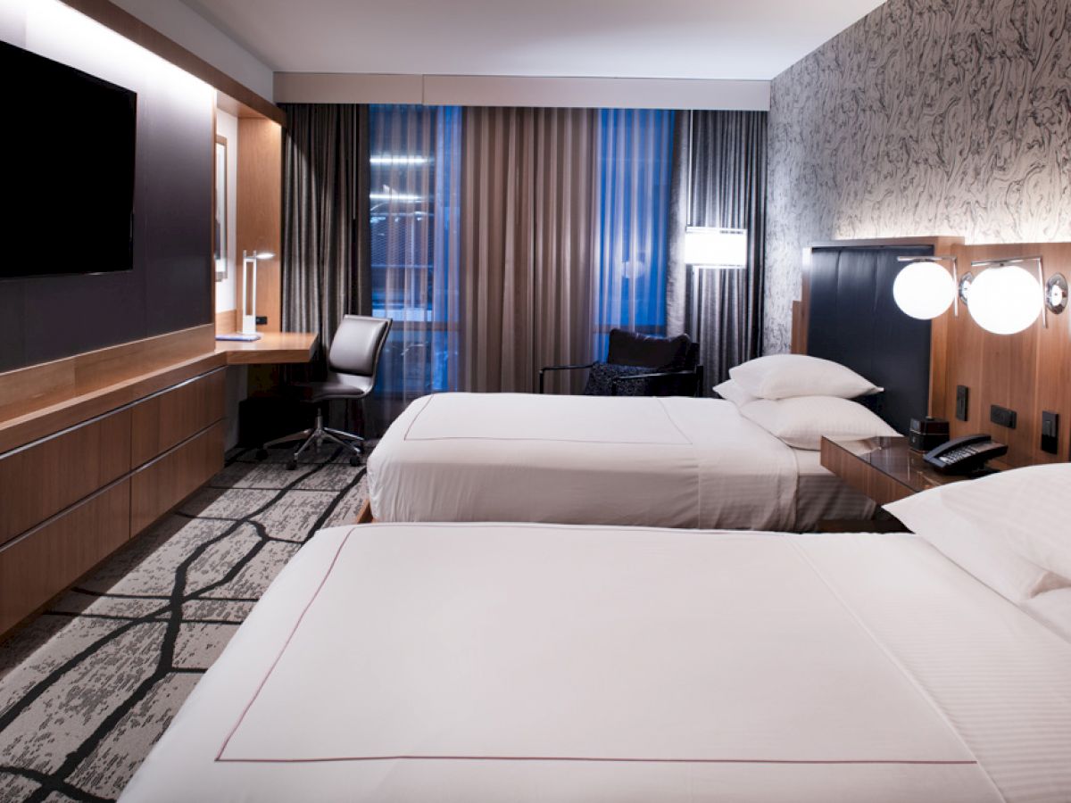 The image shows a modern hotel room with two neatly made beds, a large flat-screen TV, a desk, and contemporary decor.