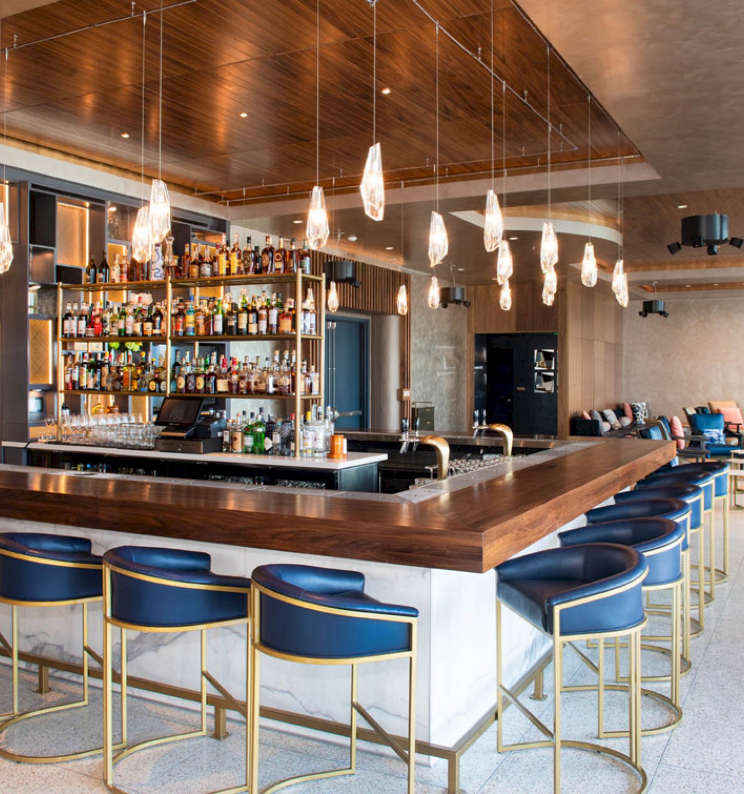 This image shows a modern bar with blue bar stools, a well-stocked liquor shelf, unique overhead lighting, and a lounge area with seating in the background.
